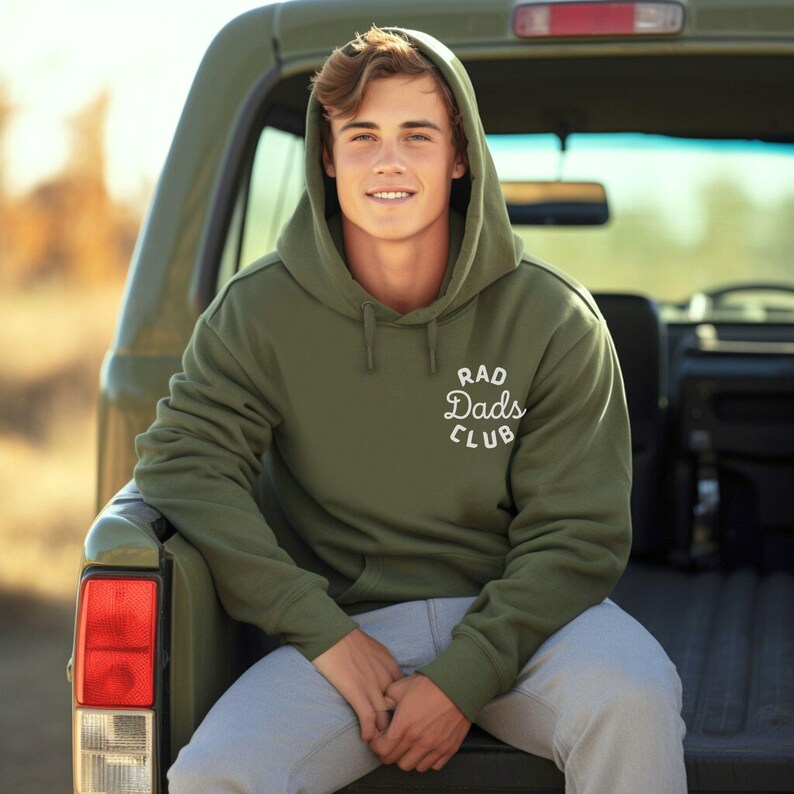 May include: A young person wearing a green hooded sweatshirt with the text "Rad Dads Club" in white. The person is sitting in the back of a pickup truck.