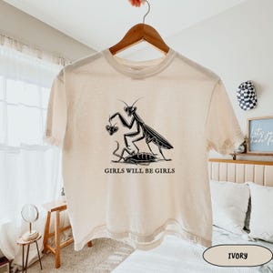 May include: Ivory-colored t-shirt with a black graphic of a praying mantis holding a heart and standing over a cockroach. The text "GIRLS WILL BE GIRLS" is below the graphic. The shirt is hanging on a wooden hanger.