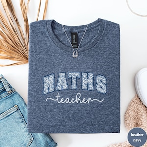 Könnte beinhalten: Marineblaues meliertes T-Shirt mit dem Schriftzug "MATHS teacher" in einem floralen Design. Das Wort "MATHS" ist in einem blauen Blumenmuster gehalten, und "teacher" ist in weißer Schrift. Eine silberne Halskette mit einem Sonnenanhänger befindet sich auf dem Shirt.