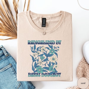 Könnte beinhalten: Ein beiges T-Shirt mit einem Blumendesign und dem Text "Radikalisiert durch grundlegende Anständigkeit".
