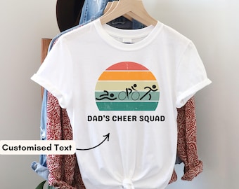 May include: White t-shirt with a colorful graphic of a swimmer, cyclist, and runner inside a sunset design. The text "DAD'S CHEER SQUAD" is below the graphic. The shirt is hanging on a wooden hanger.