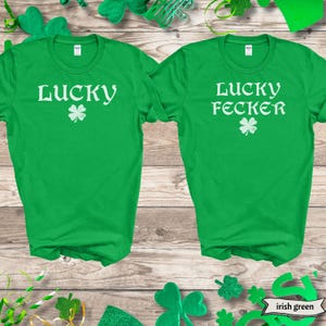 May include: Two green t-shirts with white text. The left shirt says "Lucky" with a shamrock. The right shirt says "Lucky Fecker" with a shamrock. The shirts are on a wood background.