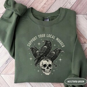 May include: Military green sweatshirt with a graphic featuring three black crows perched on a skull. The text "SUPPORT YOUR LOCAL MURDER" is arched above the crows. The sweatshirt is a solid color.