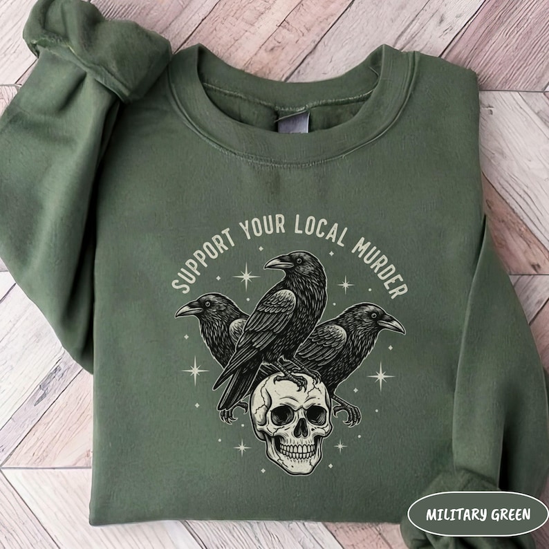 May include: Military green sweatshirt with a graphic featuring three black crows perched on a skull. The text "SUPPORT YOUR LOCAL MURDER" is arched above the crows. The sweatshirt is a solid color.