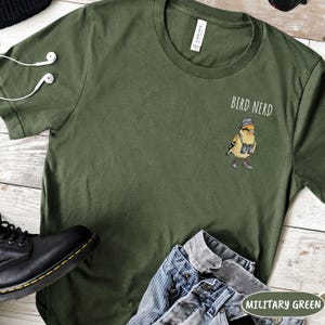 May include: Military green t-shirt featuring the words "Bird Nerd" and a cartoon bird wearing a hat and binoculars. The shirt is made of a soft material and is paired with blue jeans and black boots.