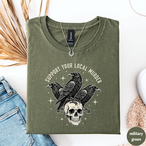 May include: Military green t-shirt with the text "SUPPORT YOUR LOCAL MURDER" encircling a graphic of three black crows perched on a skull. A silver necklace with a sun pendant is visible.