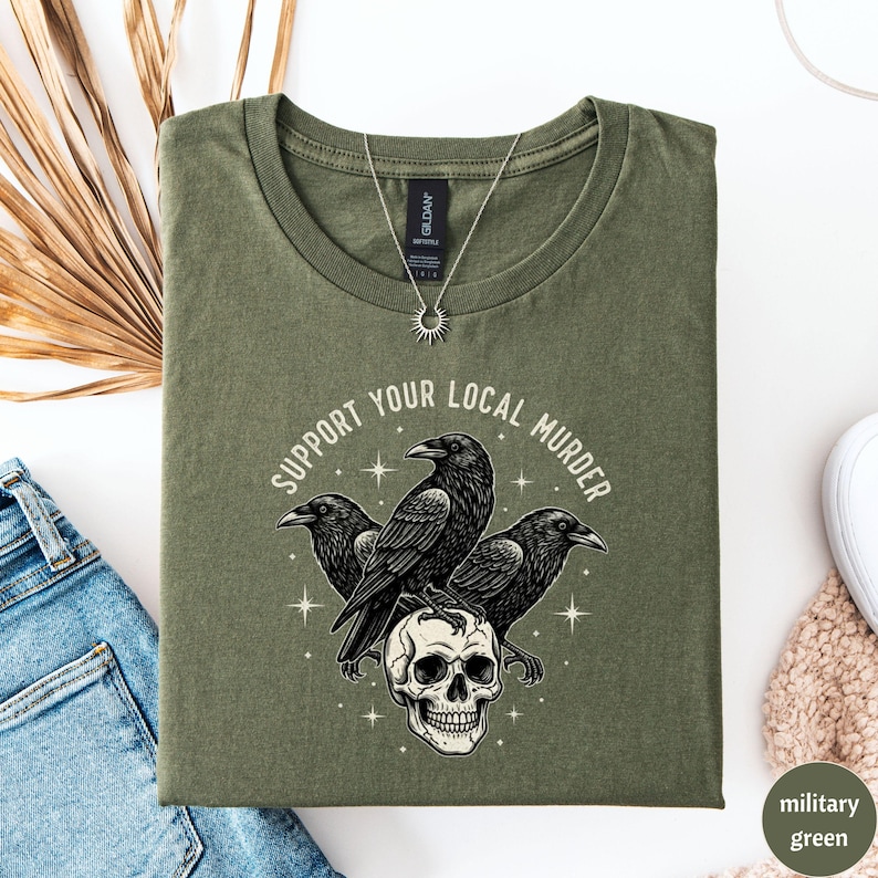 May include: Military green t-shirt with the text "SUPPORT YOUR LOCAL MURDER" encircling a graphic of three black crows perched on a skull. A silver necklace with a sun pendant is visible.