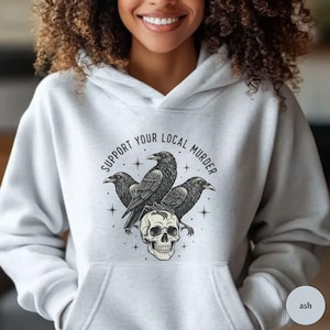 May include: Ash gray hoodie with a graphic featuring three black crows perched above a skull. The text "SUPPORT YOUR LOCAL MURDER" is printed above the crows in a circular design. The hoodie has a front pocket and a drawstring hood.