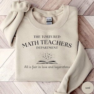 Puede incluir: Una sudadera de color arena con el texto "THE TORTURED MATH TEACHERS DEPARTMENT" y la frase "All is fair in love and logarithms". También se presenta un gráfico de un libro abierto con números radiantes.
