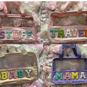 May include: Four clear plastic tote bags with different coloured fabric exteriors and colourful fuzzy letters. The bags are labelled "STUFF", "TRAVEL", "BABY", and "MAMA".