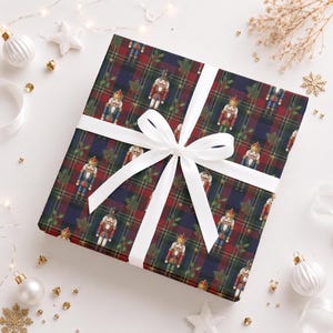 May include: A square gift box wrapped in dark blue, red, and green tartan paper featuring a repeating pattern of nutcracker figures and holly sprigs. The package is tied with a crisp white satin ribbon tied in a centred bow on top.