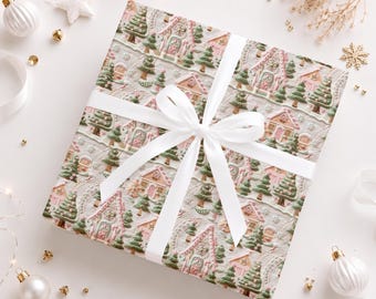 May include: A square gift box wrapped in paper featuring a repeating pattern of gingerbread houses and evergreen trees in shades of pink, green, and white. The box is tied with a smooth white satin ribbon arranged in a centered bow.
