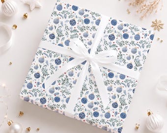 May include: A square gift box wrapped in white paper featuring a repeating pattern of blue Christmas ornaments and green pine branches, tied with a smooth white satin ribbon bow centered across the top surface of the package.