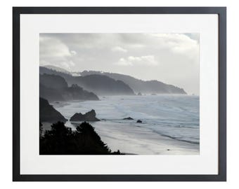 May include: A framed landscape photograph of a coastal scene. The image features a beach, ocean waves, and rolling hills under a cloudy sky. The photograph is in a black frame with a white mat.