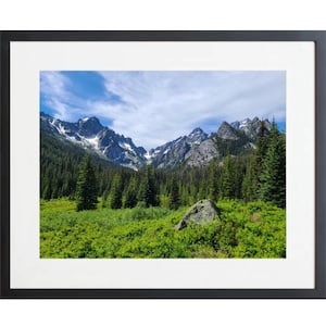 May include: A framed landscape print showing a mountain range with snow-covered peaks under a blue sky with clouds. The foreground features lush green vegetation and evergreen trees. The print is in a black frame with a white mount.