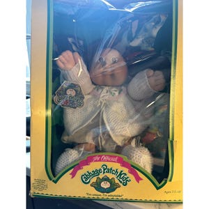 May include: A Cabbage Patch Kids doll in its original yellow and green cardboard packaging. The doll has a round face, brown hair, and wears a white knitted sweater and matching booties. The box features the Cabbage Patch Kids logo and text.