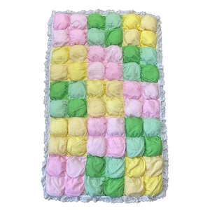 May include: A rectangular, handmade quilt featuring a patchwork design of puffy squares in pastel colors. The squares are arranged in rows and columns of pink, green, and yellow gingham and solid fabrics. The quilt is trimmed with a white lace border.