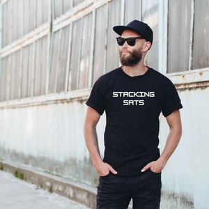 以下が含まれることがあります： 黒いTシャツを着て、白い文字で「STACKING SATS」と書かれた男性。黒いベースボールキャップとサングラスをかけています。