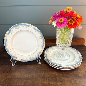 May include: Set of four white porcelain plates with blue and gold trim. The plates have a scalloped edge and a delicate floral design. The plates are stacked on top of each other.