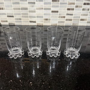 May include: A set of four clear glass tumblers arranged in a row. Each cylindrical glass features a distinct base decorated with a ring of small, raised glass spheres or bubbles encircling the bottom edge of the vessel.