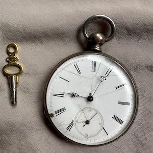 May include: A silver pocket watch with a white face and black Roman numeral hour markers. The watch has a cracked glass face and a small second hand dial. The watch is accompanied by a gold key.