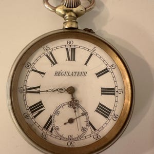 May include: A vintage pocket watch with a white face, black Roman numerals, and a smaller subdial. The watch has a gold-colored bezel and a silver-colored winding stem. The word "REGULATEUR" is printed on the face.