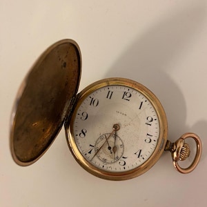 Puede incluir: Un reloj de bolsillo dorado con tapa abierta, que revela una esfera blanca con números y agujas negras. El reloj tiene un pequeño dial de segundos y un vástago de cuerda. Fondo neutro.