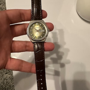 Peut inclure: Une montre argentée avec un bracelet en cuir marron. Le cadran de la montre est vert et comporte une fenêtre de date. Le nom de la marque "SANDOZ" est imprimé sur le cadran de la montre.