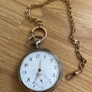 Könnte beinhalten: Eine silberne Taschenuhr mit weißem Zifferblatt und schwarzen römischen Ziffern. Die Uhr hat eine goldene Kette.