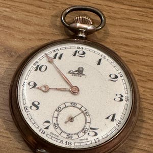 May include: A silver pocket watch with Roman numeral markings and a small sub-dial. The watch face features a lion logo with the text "LJOR" underneath. The watch has a white face and black hands.