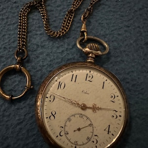 Puede incluir: Un reloj de bolsillo de plata con una esfera blanca y marcas de números romanos negros. El reloj tiene una cadena dorada unida a él. La esfera del reloj dice "Odin".