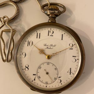 Puede incluir: Reloj de bolsillo antiguo con esfera blanca, números negros y manecillas doradas. El reloj tiene una esfera secundaria y está sujeto a una cadena plateada. La esfera del reloj dice "Gust. Dahl Malmö".