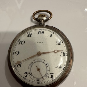 Puede incluir: Reloj de bolsillo de plata con una esfera blanca y marcas de números romanos negros. El reloj tiene una corona dorada y una aguja de segundos negra. La esfera del reloj también tiene un círculo más pequeño con marcas negras para los segundos. La esfera del reloj dice "EDEN" en la parte superior.