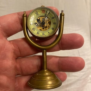 Pode incluir: Um relógio de latão com uma cúpula de vidro transparente e um mecanismo de engrenagem visível. O relógio está em um suporte decorativo com uma base redonda.