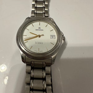 May include: A silver Grovana automatic watch with a white face, gold hands, and a date window. The watch has a silver metal band.