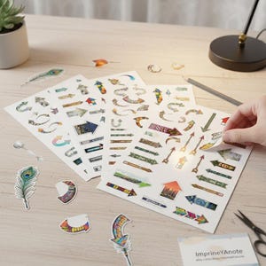 Puede incluir: Hojas de pegatinas con temática de flechas en varios colores y diseños, incluyendo flechas rectas, curvas y decorativas. Algunas pegatinas están sueltas, junto con una pluma de pavo real y una cuchara. Las pegatinas están sobre una superficie de madera.