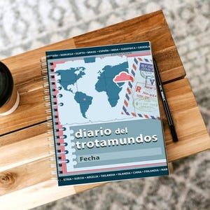 Puede incluir: Un cuaderno de espiral con un diseño de mapa del mundo y el texto "diario del trotamundos". Un bolígrafo negro y una taza de café están sobre una superficie de madera. El cuaderno tiene una cubierta azul claro.