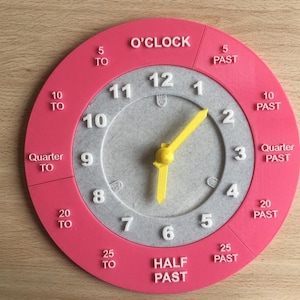 May include: A pink and gray circular clock face with yellow hands. The clock face has numbers 1 through 12 and markings for 5, 10, 15, 20, 25, and 30 minutes past and to the hour. The clock face also has the words "O'CLOCK", "HALF PAST", "QUARTER TO", and "QUARTER PAST".