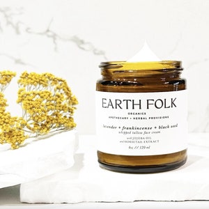 Puede incluir: Un tarro de vidrio de crema facial de sebo batido Earth Folk Organics con lavanda, incienso y semillas negras. El tarro es de 4 oz / 120 ml y está sobre una superficie blanca.