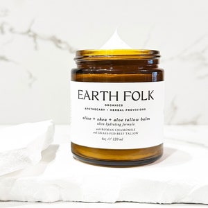 Puede incluir: Un tarro de vidrio marrón con una etiqueta blanca que dice "EARTH FOLK ORGANICS APOTHECARY HERBAL PROVISIONS olive + shea + aloe tallow balm ultra hydrating formula with ROMAN CHAMOMILE and GRASS-FED BEEF TALLOW 4oz // 120 ml". El tarro está lleno de una crema blanca.