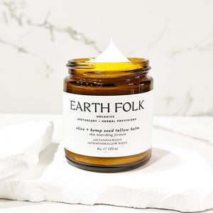 May include: A brown glass jar with a white label that reads "EARTH FOLK ORGANICS APOTHECARY HERBAL PROVISIONS olive + hemp seed tallow balm skin nourishing formula with SANDALWOOD and MARSHMALLOW ROOT 4oz / 120 ml". The jar is filled with a white cream.