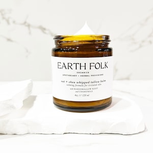 May include: A brown glass jar with a white label that reads "EARTH FOLK ORGANICS APOTHECARY HERBAL PROVISIONS oat + shea whipped tallow balm calming formula for irritated skin with MARSHMALLOW ROOT and CHAMOMILE 4oz // 120 ml". The jar contains a white cream.