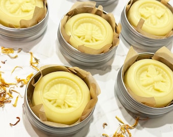 May include: Six small silver tins with lids, each containing a round, pale yellow balm with a bee design embossed on the top. The tins are arranged in a semi-circle on a white surface with scattered yellow flower petals.