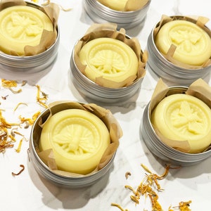 Puede incluir: Seis pequeñas latas de plata con tapas, cada una contiene un bálsamo redondo de color amarillo pálido con un diseño de abeja en relieve en la parte superior. Las latas están dispuestas en semicírculo sobre una superficie blanca con pétalos de flores amarillas esparcidos.