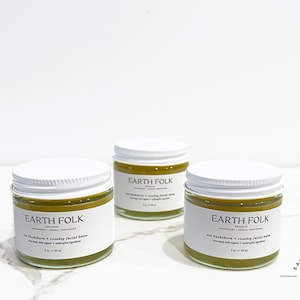 May include: Three small jars of green facial balm with white lids and labels that read "EARTH FOLK" in black text. The jars are on a white marble surface.