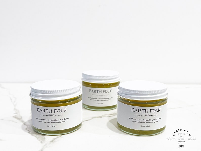 May include: Three small jars of green facial balm with white lids and labels that read "EARTH FOLK" in black text. The jars are on a white marble surface.