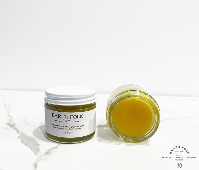 May include: Two glass jars of yellow facial balm with white lids. The jar on the left has a white label with the text "EARTH FOLK" and "Sea Buckthorn & Rosehip Facial Balm". The jar on the right is open, showing the yellow balm inside.