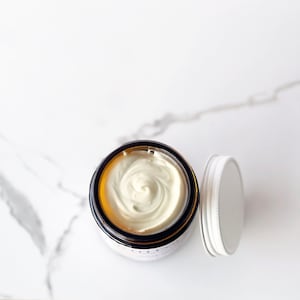 May include: A white cream in a glass jar with a brown lid. The jar is on a white marble surface.