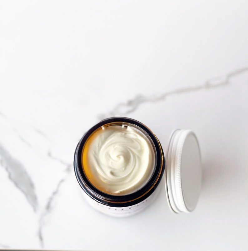 May include: A white cream in a glass jar with a brown lid. The jar is on a white marble surface.