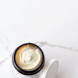 May include: A close-up of a white cream in a glass jar with a brown lid. The cream is swirled and has a smooth texture.